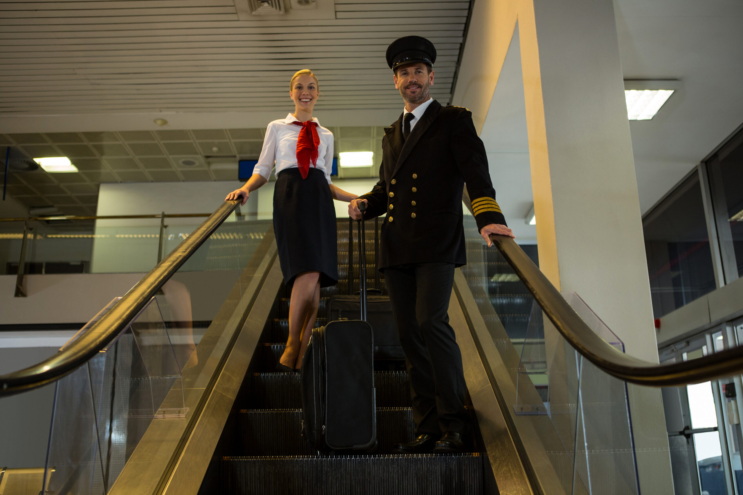 Pilot and air hostess with their trolley bags standing on escalator in the airport terminal